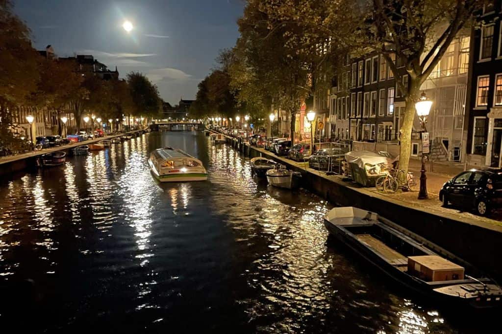 A canal boat sailing away in the evening under a moonlit sky in Amsterdam.