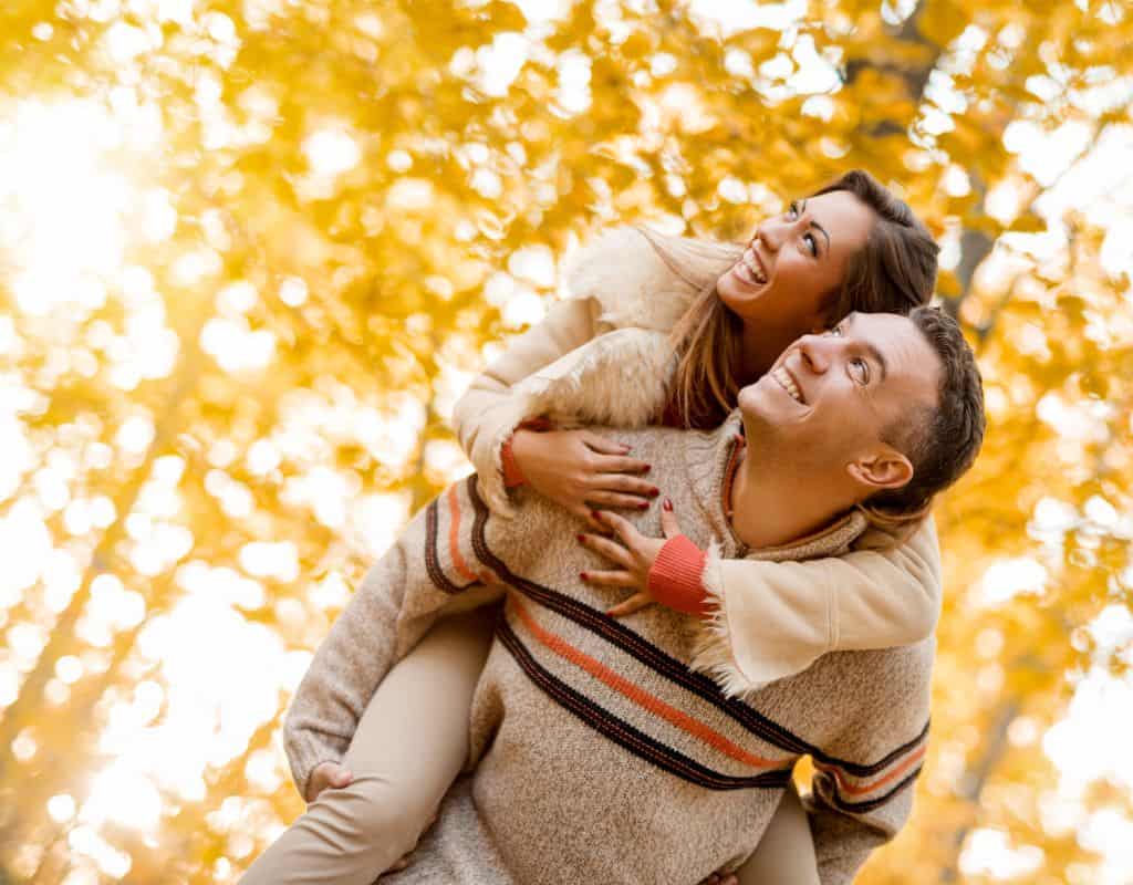 A woman riding piggy back on her boyfriends back as they both look up towards the sky with yellow trees in the background on a sunny fall afternoon.