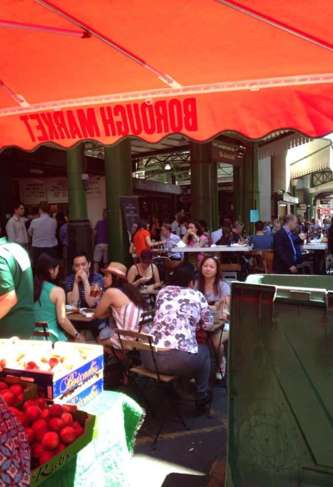 What is London Famous For People sitting in the central sitting area at the Borough Market as seen from under a fruit vendor's awning.