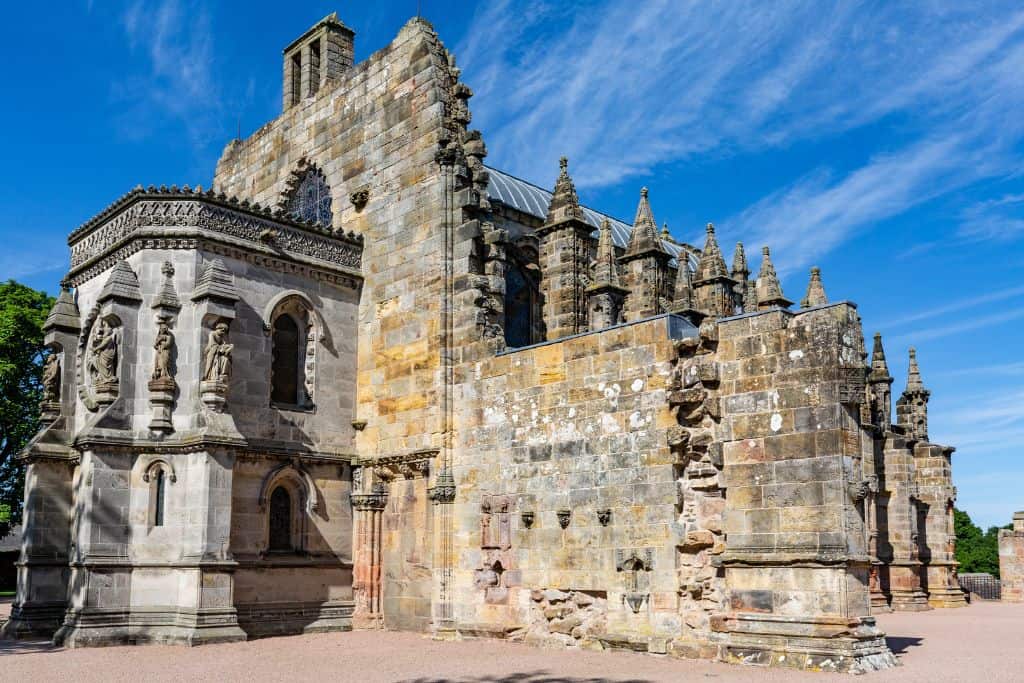 Day Trips From Edinburgh Without a Car The rocky exterior of Rosslyn Chapel, with many turrets and statues adorning the outside.