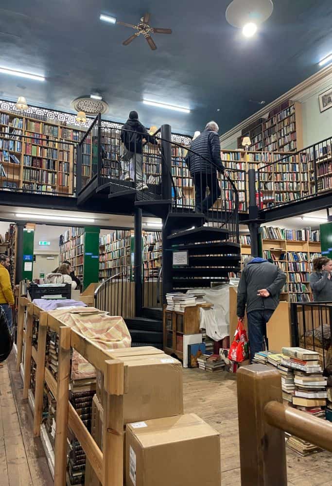 Scotland Souvenirs Interior of a bookstore with tall bookshelves filled with books and a spiral staircase, with people browsing the collection on different levels.