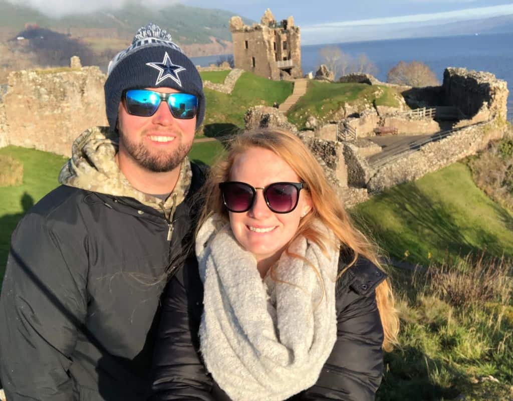 Scotland Souvenirs Taylor and her husband standing in front of the ruins of Urquhart Castle overlooking Loch Ness, both smiling and wearing sunglasses, with a scenic background of the lake and hills.