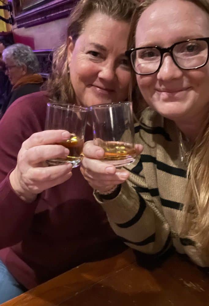 Shops-on-Victoria-Street-Edinburgh Taylor and a girlfriend smiling and holding up glasses of whisky in a dimly lit pub. The backdrop features other patrons and a cozy, relaxed atmosphere typical of a Scottish bar.