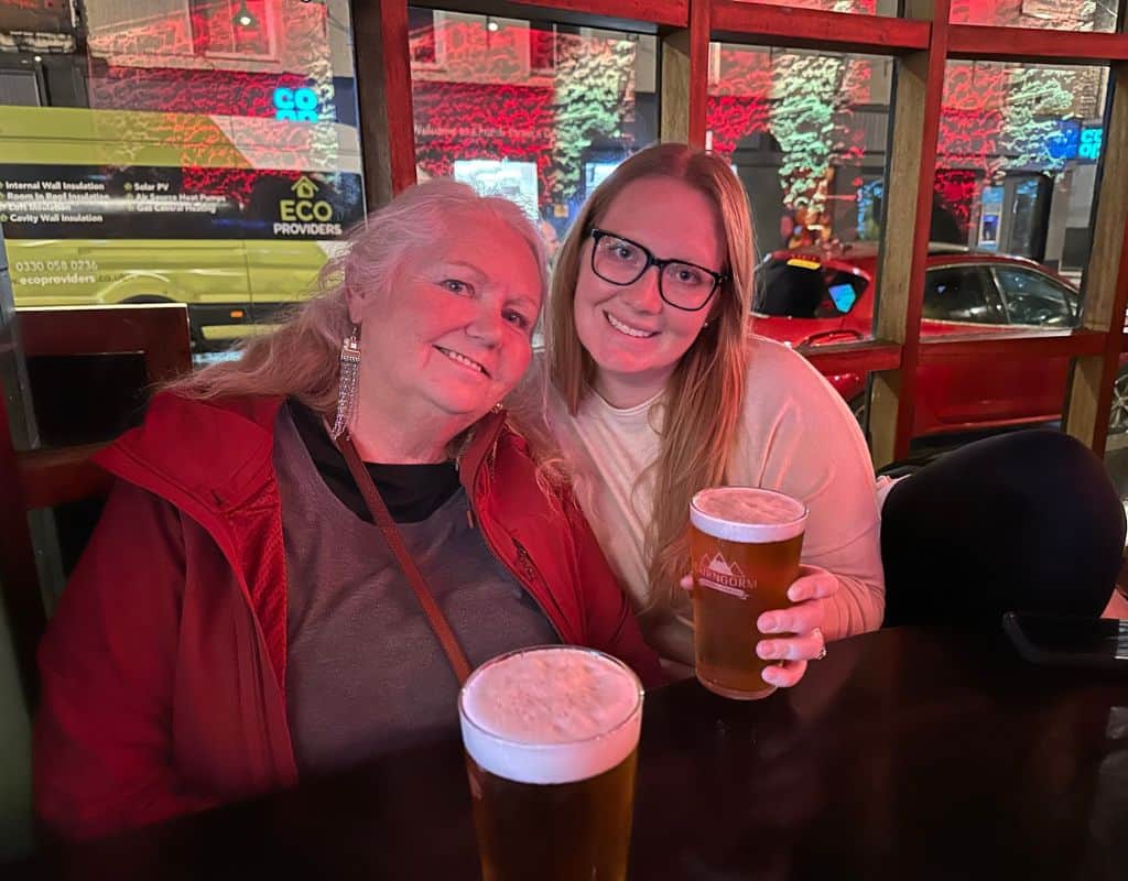 Taylor and a girlfriend sitting at a pub, each holding a pint of beer, smiling and enjoying the cozy, casual atmosphere.