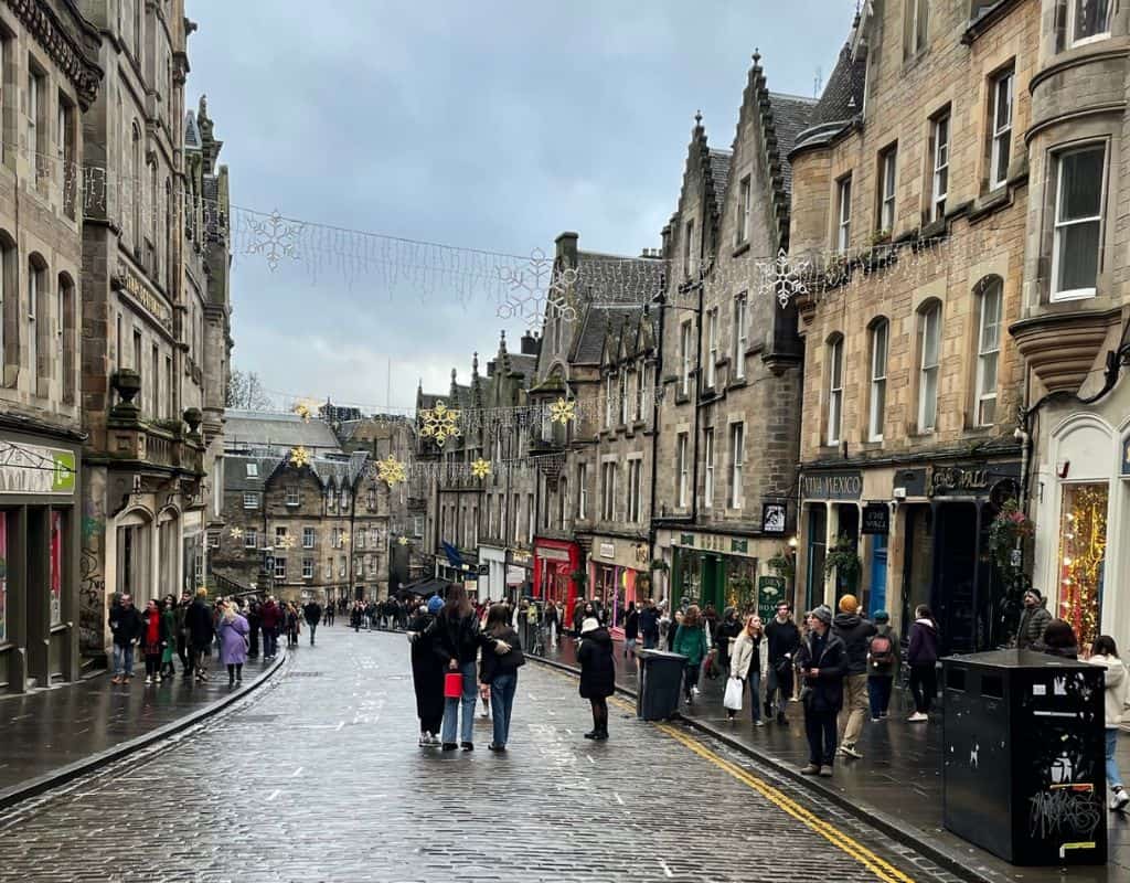 A lively street in Edinburgh filled with people walking along the cobblestone road, with festive holiday lights strung above the historic stone buildings.