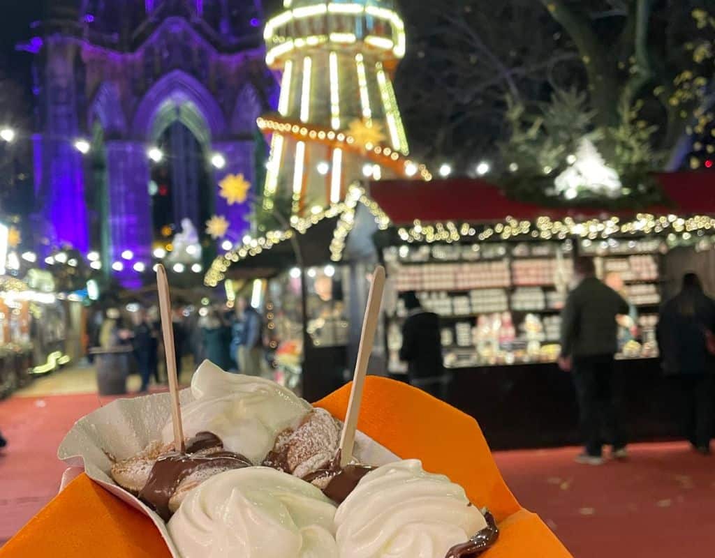 A close-up of a serving of mini pancakes topped with whipped cream and drizzled with chocolate sauce, held up at a bustling outdoor Christmas market in Edinburgh. Festive lights and a carousel are visible in the background.