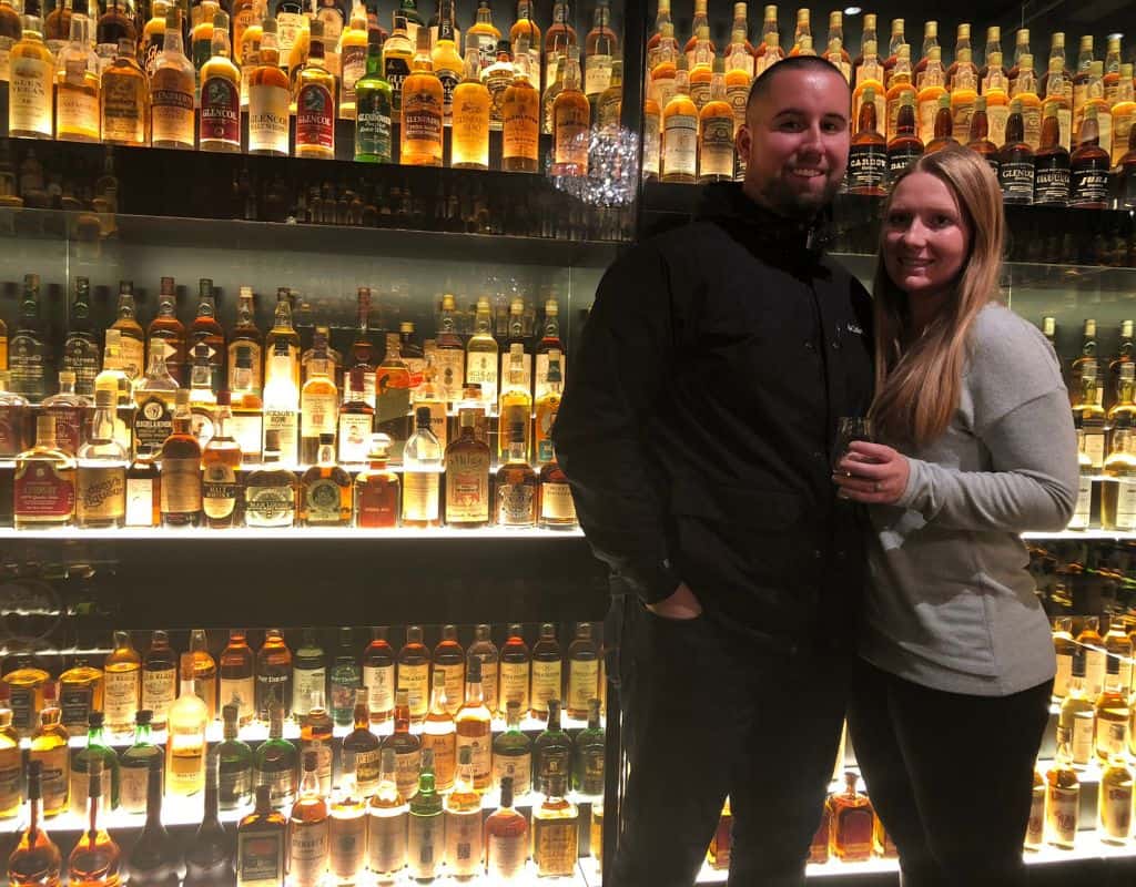 Taylor and her partner smiling and posing in front of a large collection of whisky bottles in Edinburgh. The bottles are arranged in illuminated shelves, showcasing a wide variety of Scotch whisky.
