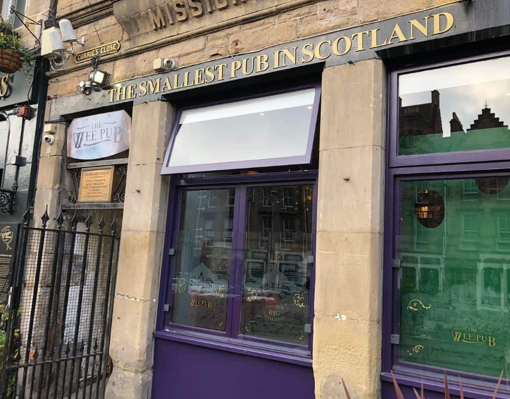 The exterior of "The Wee Pub," which claims to be the smallest pub in Scotland. The sign above the purple storefront proudly displays its name, with a small side street and a black iron gate visible on the left.