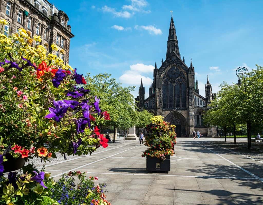 One Week Scotland Itinerary Glasgow Cathedral stands tall with its pointed spires and large stained-glass windows, framed by blooming flowers and leafy trees—a beautiful stop for travelers exploring a 1 week Scotland itinerary.