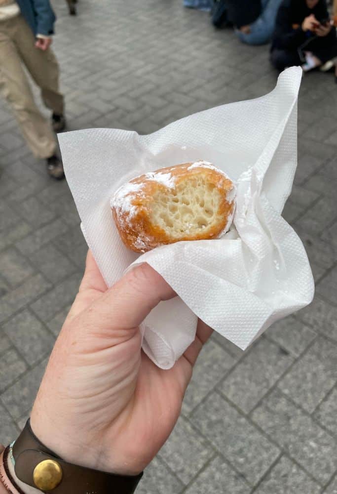 A hand holding a half-eaten Dutch oliebol, a deep-fried doughnut dusted with powdered sugar, wrapped in a white napkin while walking through a busy street.