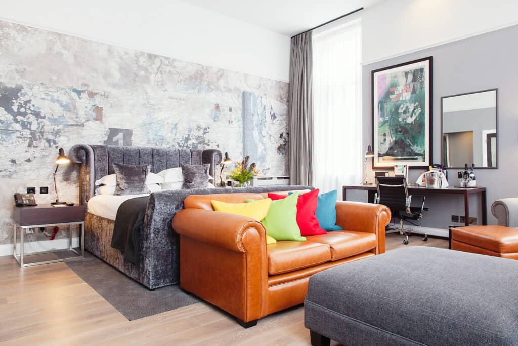 Historic Hotels in Edinburgh A bright and airy hotel room with geographic wallpaper behind a large grey fabric bed. A leather sofa and a desk adorn the rest of the suite with floor length white curtains in the background.