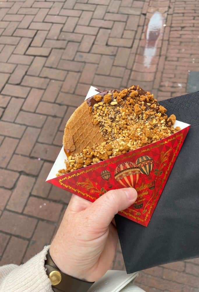 A hand holding a Dutch stroopwafel covered in chocolate and crushed nuts, served in a festive red paper sleeve with a cobblestone street in the background.