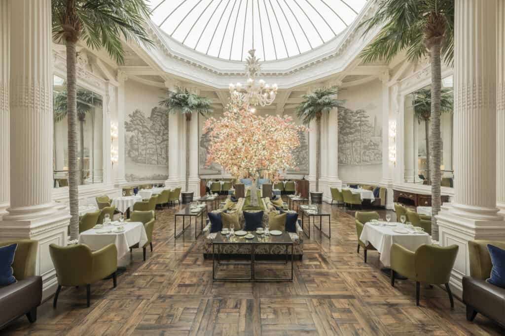 Historic Hotels in Edinburgh A large ballroom type room with white paneling and wood flooring with palm trees along the sides. The room is clearly historic and is set with small tables and green plush chairs for tea at the historic hotel in Edinburgh, the Balmoral.