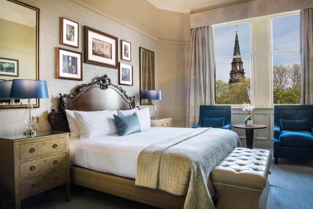 Historic Hotels in Edinburgh A photo of a hotel room with a large bed and historic decorations in navy blue. The Sir Walter Scott Monument can be seen through the window, as the suite is named for Sir walter Scott in one of the most historic hotels in Edinburgh.