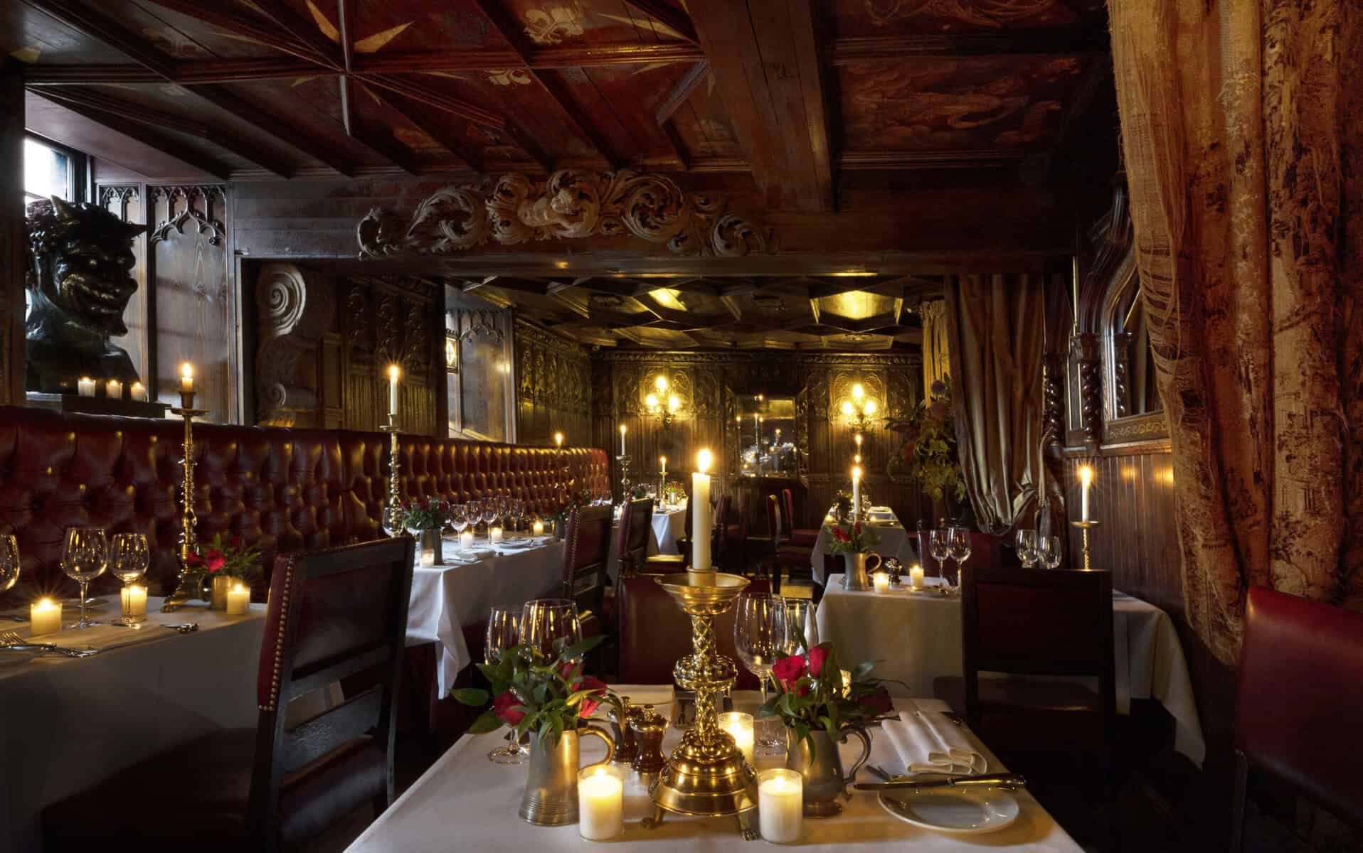 A restaurant in The Witchery, a historic hotel in Edinburgh, with white-linen tablecloth set tables. Gold candlesticks are lit on the tables, with red leather setees and chairs as seating. The brown wood ceiling is elegantly decorated in period art.