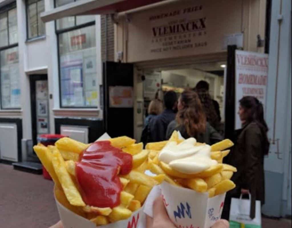 Two paper cones filled with fries, one topped with ketchup and the other with mayonnaise, held in front of Vleminckx, a famous fries stand in Amsterdam with a small queue of customers. Vleminckx serves some of the best French fries in Amsterdam, known for their crispy texture and rich sauces.