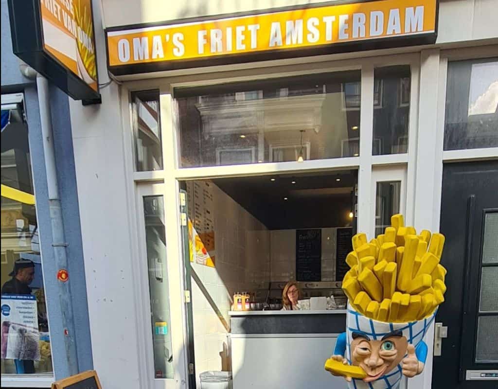 The exterior of Oma’s Friet Amsterdam, a popular fries shop, featuring a bright yellow sign and a playful fries mascot statue in front of the entrance. Oma’s Friet is a go-to spot in Amsterdam for classic Dutch fries with homemade sauces.