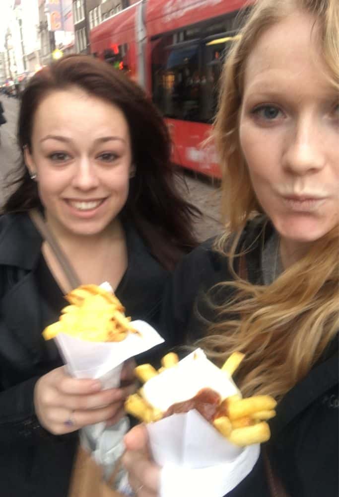 Taylor and a friend holding paper cones filled with golden fries topped with sauce while walking on a busy Amsterdam street, with a red tram passing behind them. A delicious street food experience in the heart of Amsterdam.