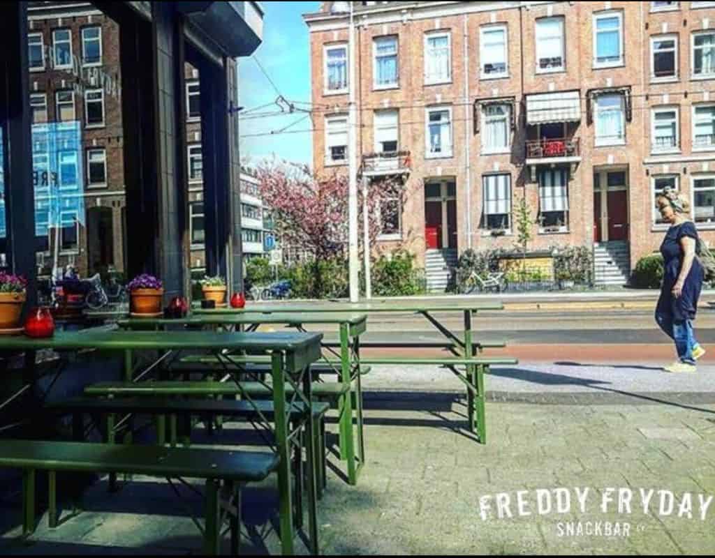 An outdoor seating area at Freddy Fryday Snackbar in Amsterdam, featuring green picnic tables and a view of traditional Dutch townhouses. A great place to enjoy French Fries in Amsterdam with a scenic street view.