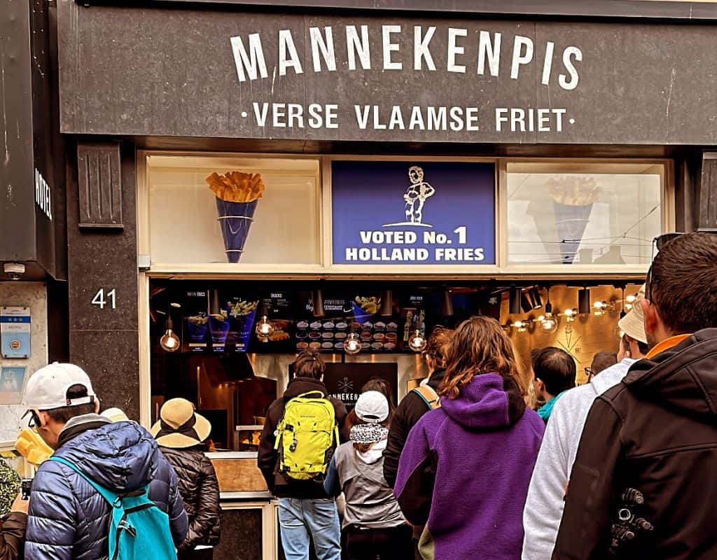 A long queue of people waiting outside Manneken Pis, a famous fries shop in Amsterdam, with a sign that reads "Voted No. 1 Holland Fries." Manneken Pis is an iconic spot in Amsterdam for thick, crispy French fries with a variety of sauces.