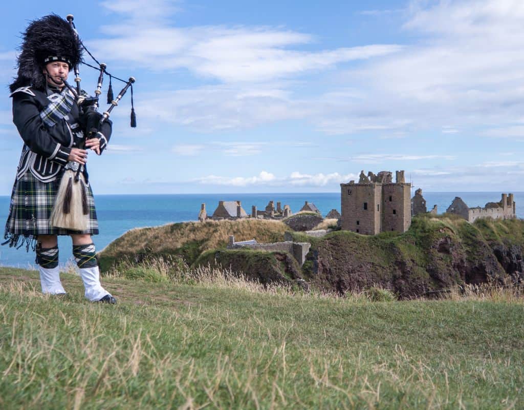 A man dressed in full traditional Scottish Highland attire plays the bagpipes on a grassy cliffside, with the ruins of a medieval stone castle and the blue ocean in the background.