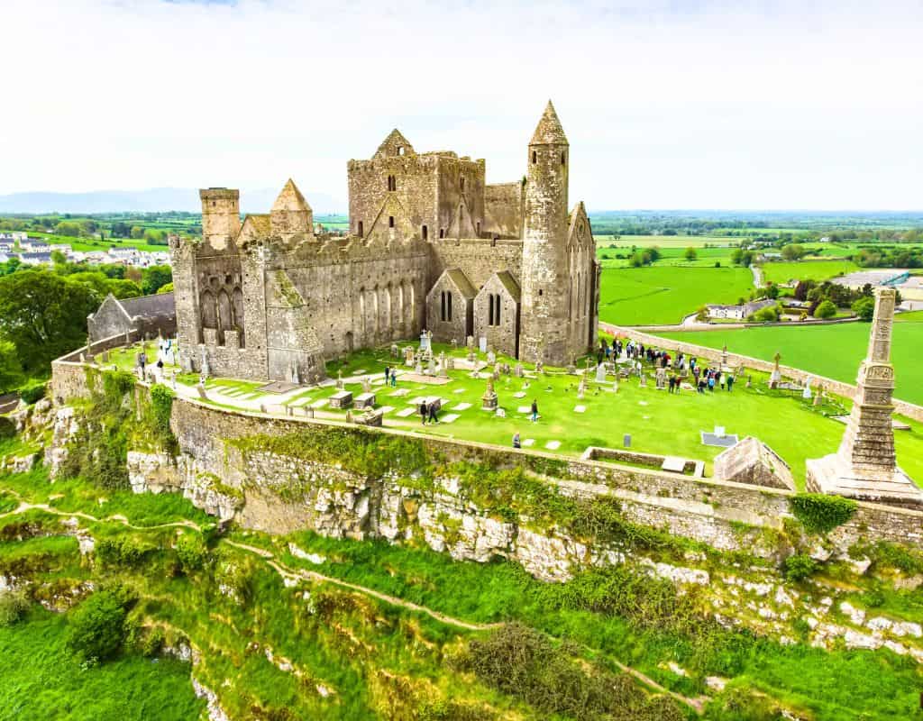 A scenic landscape shows the Rock of Cashel in Ireland, an ancient castle-like cathedral perched on a limestone hill surrounded by a cemetery and lush green fields.