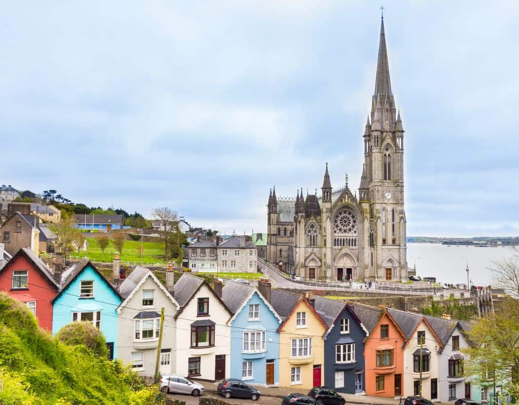 Colorful row houses line a hilly street leading toward a grand Gothic cathedral with a tall spire near the waterfront in Cobh, Ireland.
