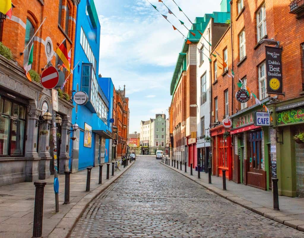A cobblestone street in Dublin's Temple Bar district is lined with pubs, cafés, and flags from various countries, with a bright blue building contrasting the traditional red brick facades.