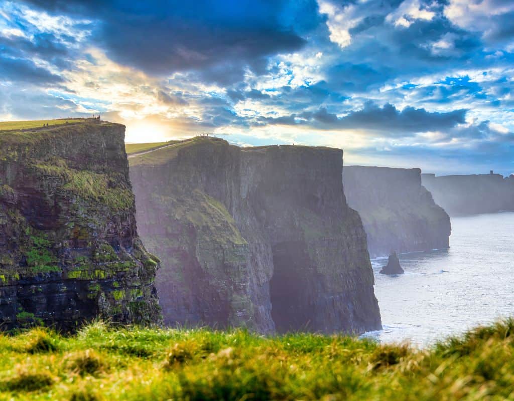 A stunning view of the Cliffs of Moher during sunset, where sunlight breaks through dramatic clouds, illuminating the jagged cliffs that rise sharply from the Atlantic Ocean.
