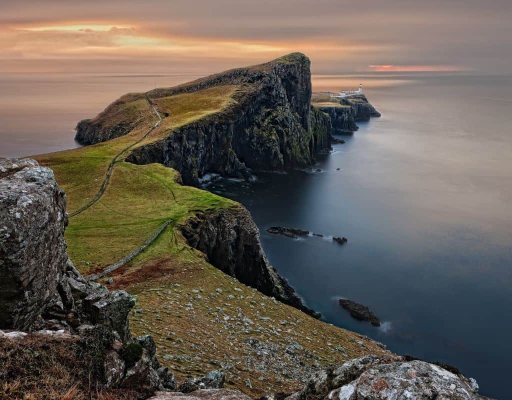 A scenic coastal headland on the Isle of Skye, Scotland, ends in a steep cliff with a lighthouse at its tip, surrounded by calm sea under a glowing twilight sky. This breathtaking view leaves some asking, “Is Scotland in Ireland?”—a common geographic mix-up.