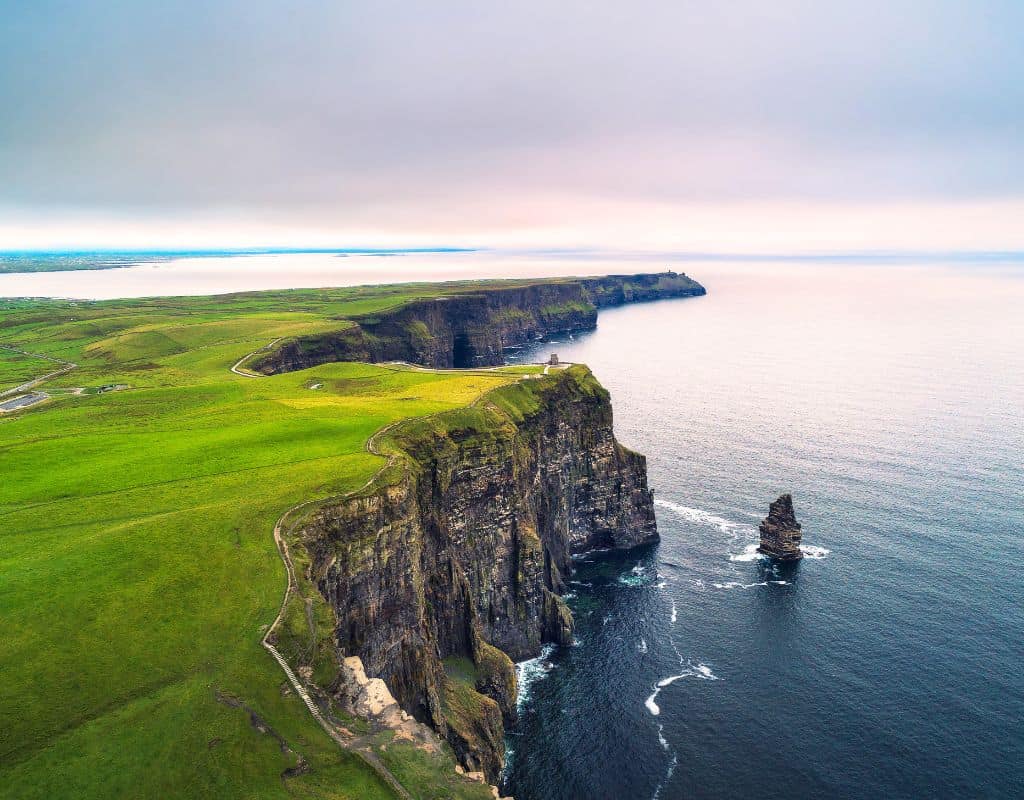 The dramatic Cliffs of Moher in Ireland stretch along the Atlantic Ocean under a soft cloudy sky, with steep drops and layered cliff faces visible from the high vantage point.