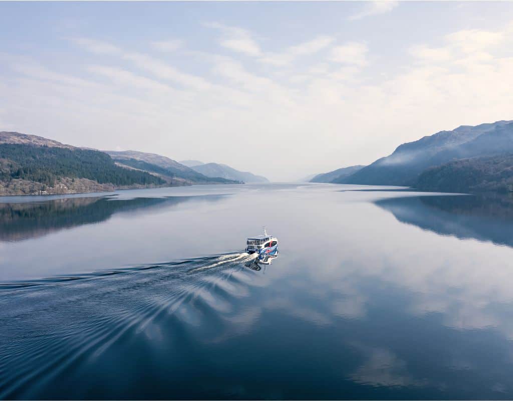 A scenic cruise boat sails across the glassy surface of Loch Ness on a calm, overcast day. Surrounded by tranquil hills and mirrored skies, the peaceful setting might leave visitors wondering, is Loch Ness worth visiting? The answer feels like a quiet yes.