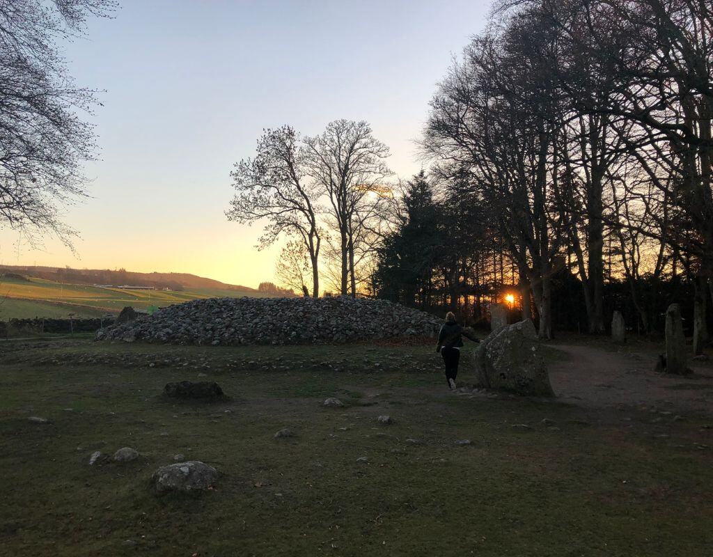 Taylor walks toward Clava Cairns at sunset, with bare trees framing the ancient burial site, casting long shadows across the grass.