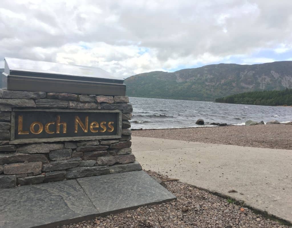 A stone sign engraved with โLoch Nessโ in gold letters sits beside the lochโs edge, with rippling dark water and forested hills in the distance beneath a cloudy sky.