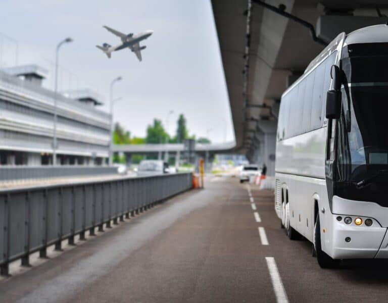 A white airport shuttle bus parked near a terminal road as an airplane takes off overhead; a typical pickup area for guests staying at Amsterdam Airport Hotels with Free Shuttle services.