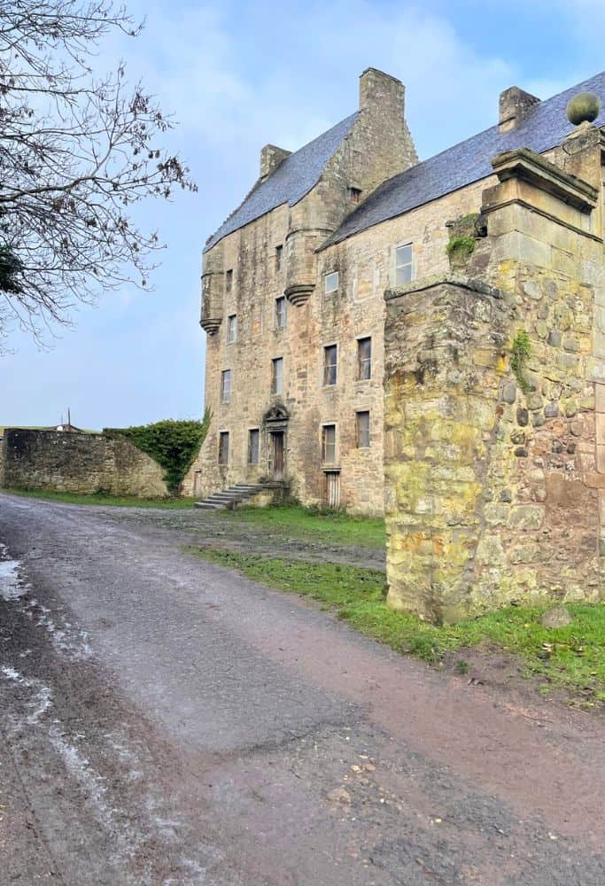 The stone tower house of Midhope Castle with its medieval architecture and weathered stone walls, located along a muddy country road under a blue sky.