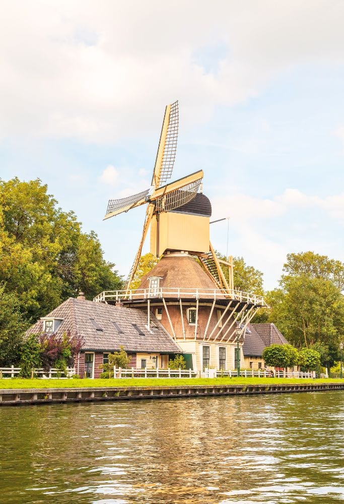 A classic Dutch windmill with a large thatched base and extended blades stands beside a canal, nestled in the peaceful Amsterdam countryside.