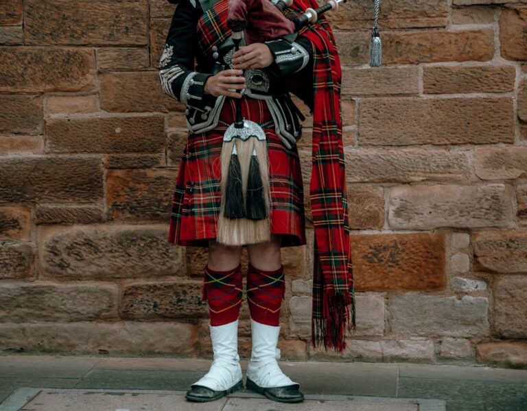 A bagpiper in full regalia, including a red tartan kilt and tall white hose, stands against a stone wall holding his bagpipes.