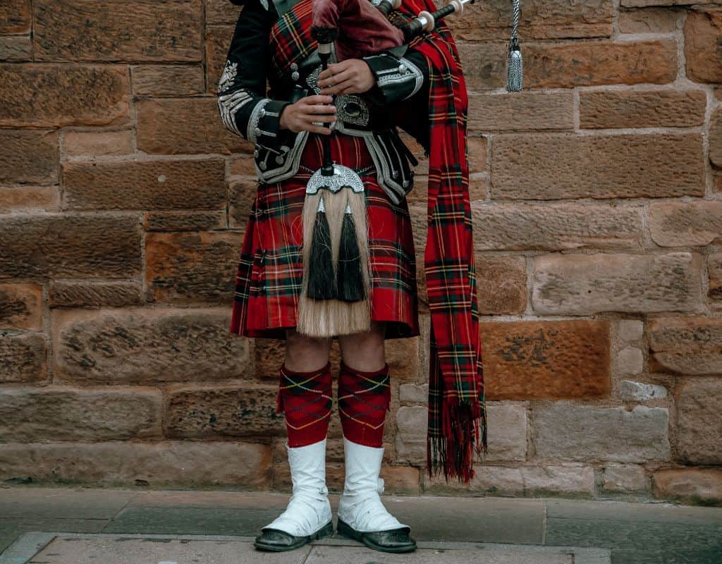 A bagpiper in full regalia, including a red tartan kilt and tall white hose, stands against a stone wall holding his bagpipes.