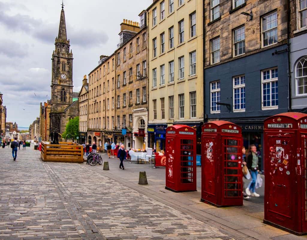 A cobbled street in the heart of Edinburgh’s Old Town features classic architecture, iconic red phone booths, and a towering clock church—right in the midst of some of the Best Places to Stay in Edinburgh.