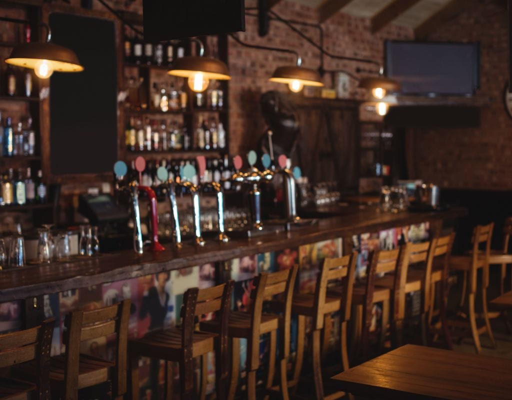 Interior of a traditional pub with a long wooden bar, high stools, hanging pendant lights, and rows of beer taps against exposed brick walls.