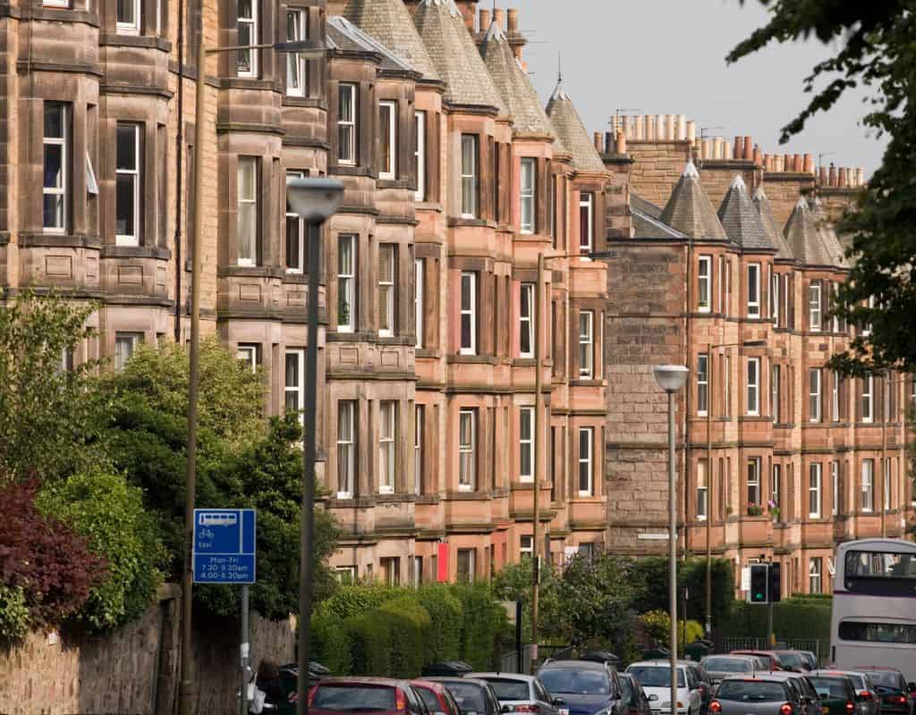 A quiet residential street in Edinburgh lined with rows of historic sandstone tenement buildings, with parked cars and leafy greenery adding to the charm of the neighborhood.