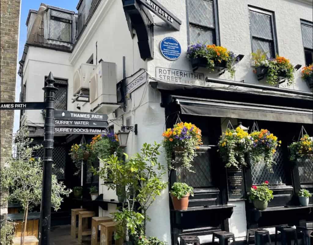Exterior of a charming white corner pub in Rotherhithe with black trim, hanging flower baskets, street signs, and outdoor stools beneath the windows.