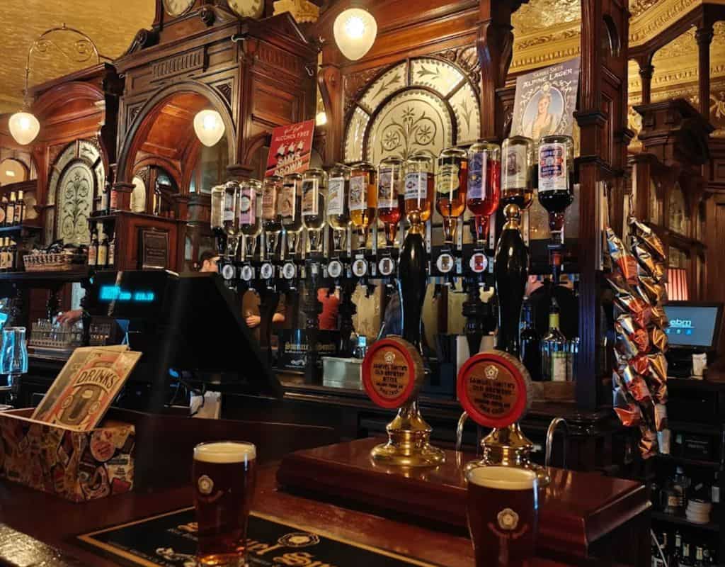 Ornate wooden pub bar lined with rows of spirits and hand-pulled ales, decorative mirrors, and two freshly poured pints representing London Oldest Pubs