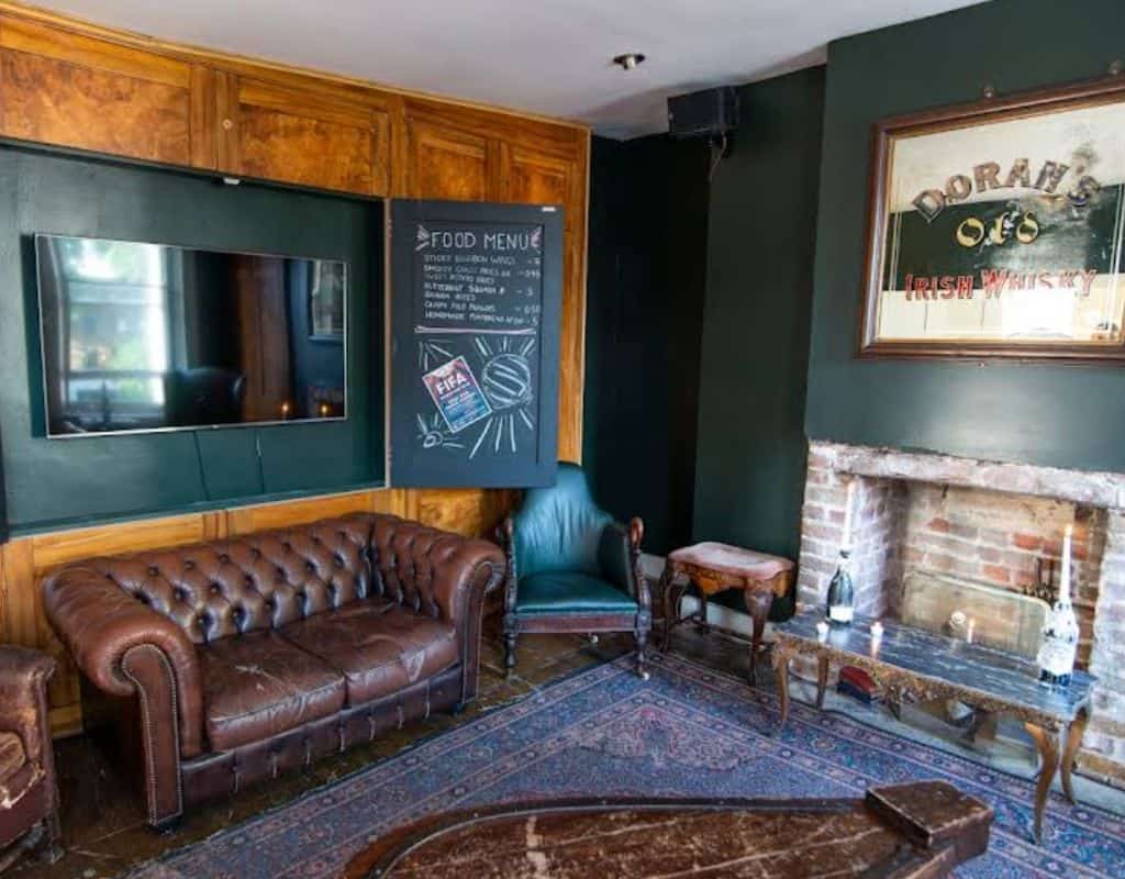 Comfortable lounge area with a brown leather Chesterfield sofa, brick fireplace, framed Irish whiskey poster, and chalkboard food menu on deep green walls.