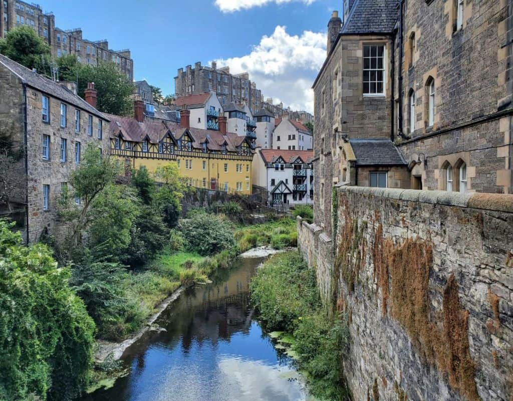 Colorful historic cottages and stone buildings line a narrow stream in the picturesque Dean Village area of Edinburgh, surrounded by lush greenery and layered city views.