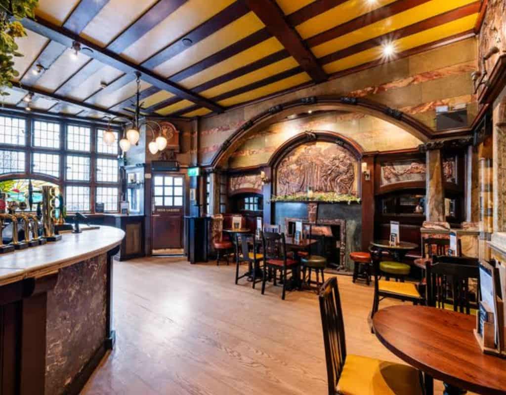 Spacious historic pub interior with carved wooden arches, detailed ceiling panels, marble columns, and classic round tables showcasing the grandeur of London Oldest Pubs.