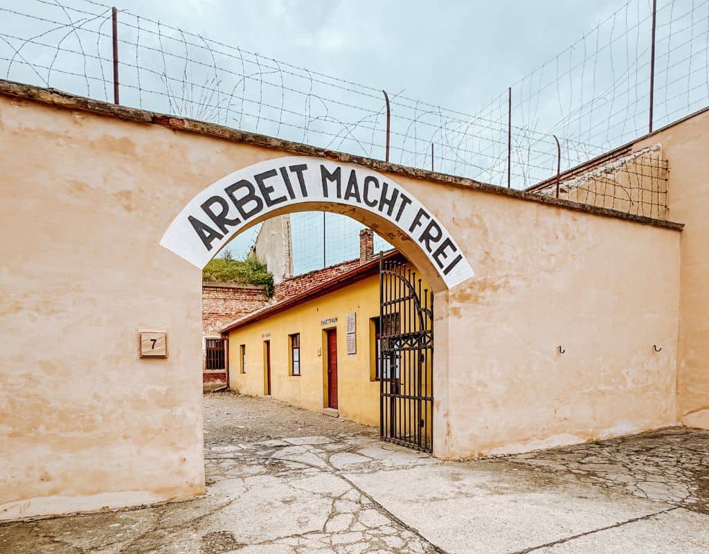 Entrance gate with the “Arbeit Macht Frei” sign at the former Theresienstadt concentration camp memorial, featuring historic prison buildings, barbed wire fencing, and a solemn courtyard near Prague.