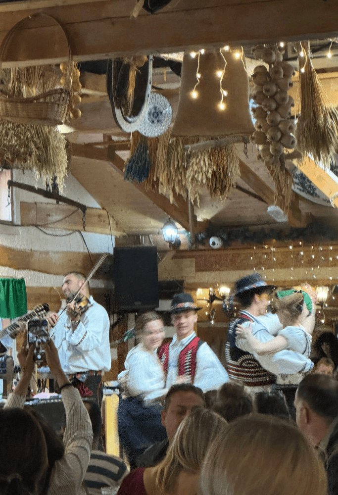Folk dancers in traditional Czech clothing performing alongside live musicians inside a rustic restaurant with warm lighting and hanging décor.