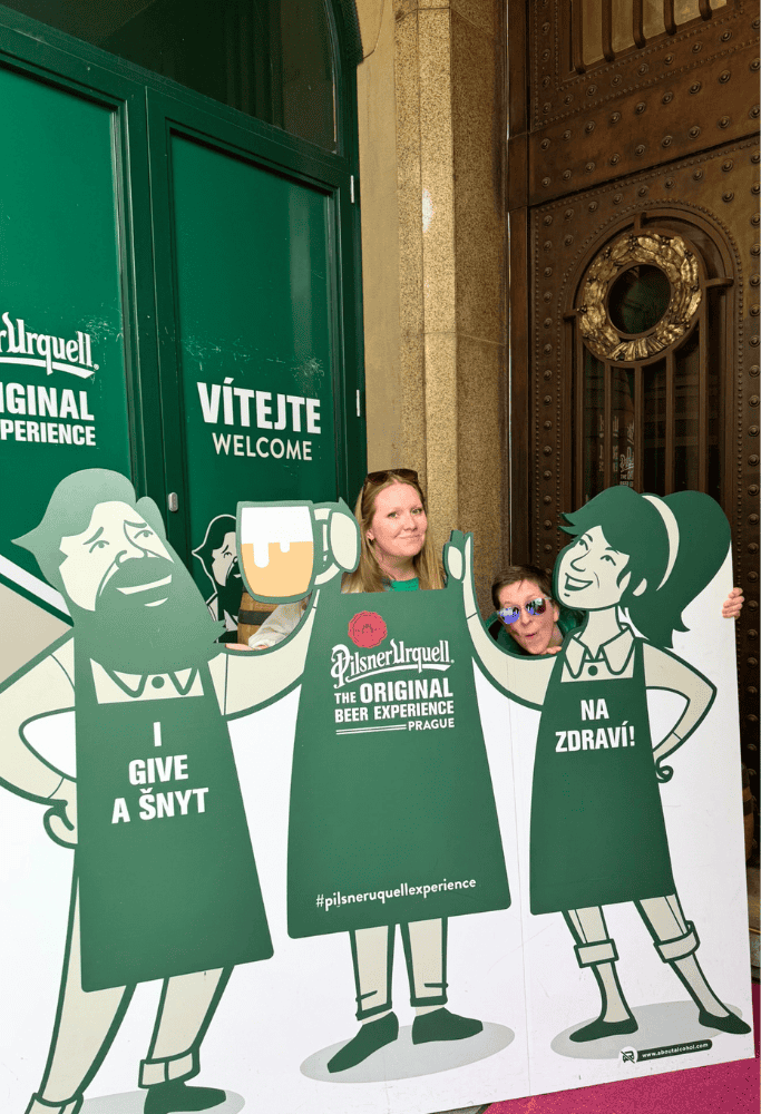 Taylor posing at the Pilsner Urquell Beer Experience entrance in Prague beside playful beer-themed displays and brewery branding.